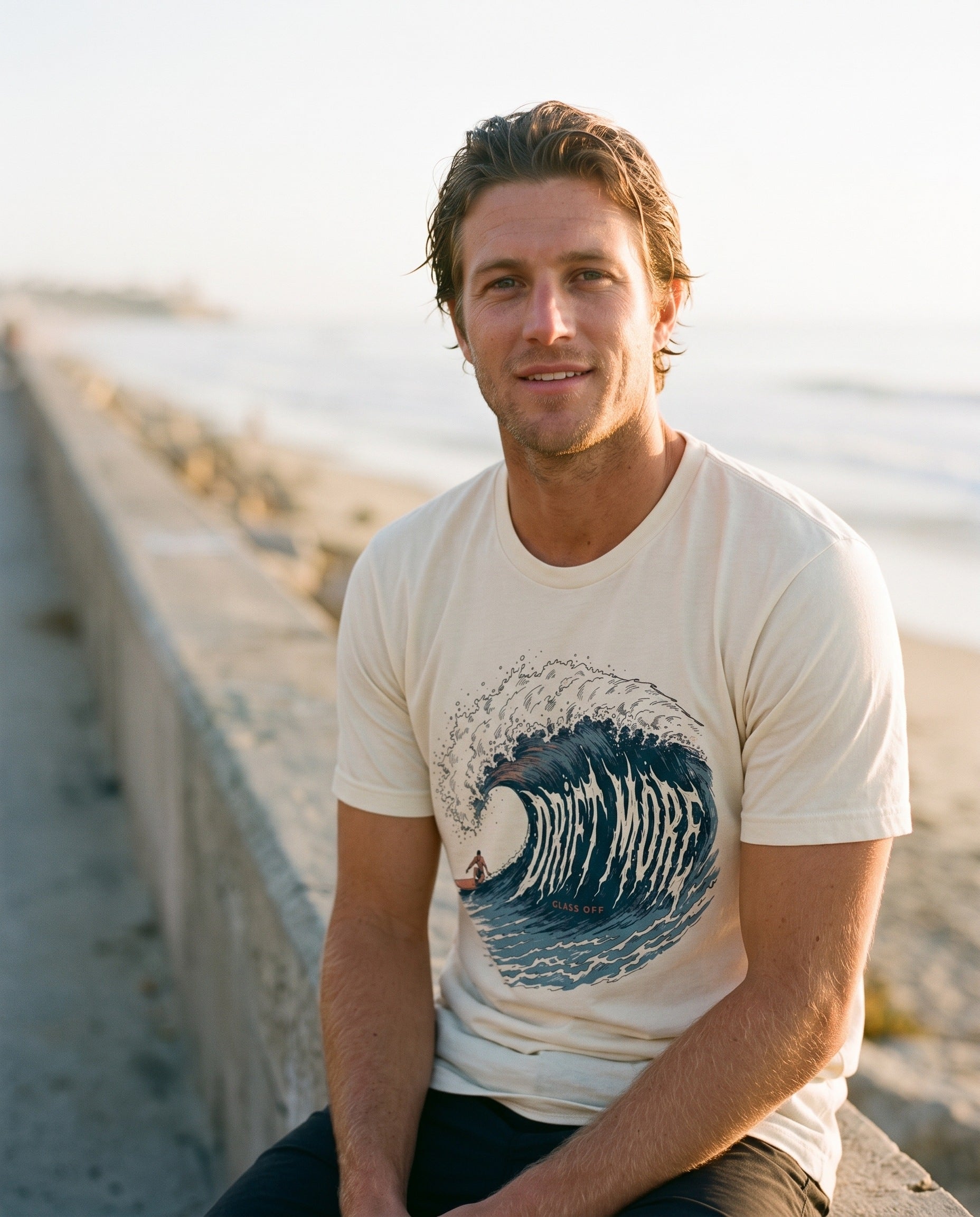 Drift More Wave Tee in natural cream, worn on a seawall at last light