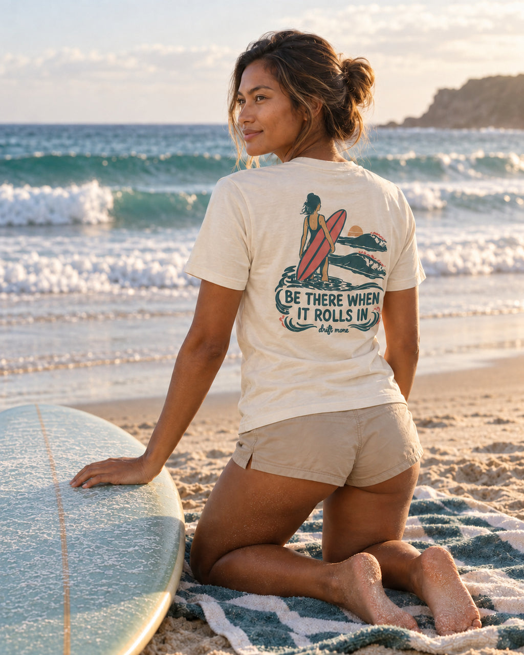 Kai wearing the Natural Be There When It Rolls In women’s tee, showing the back print beside a surfboard on the beach